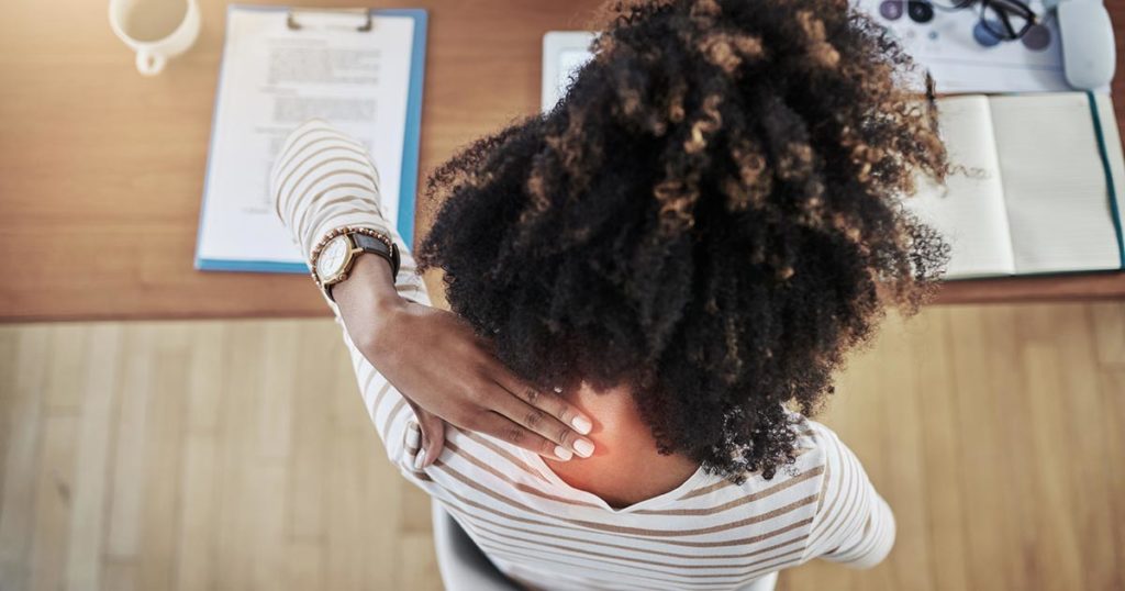 Aerial view of Black woman working at her desk, she is reaching over to the back of her neck and rubbing her itch from eczema.