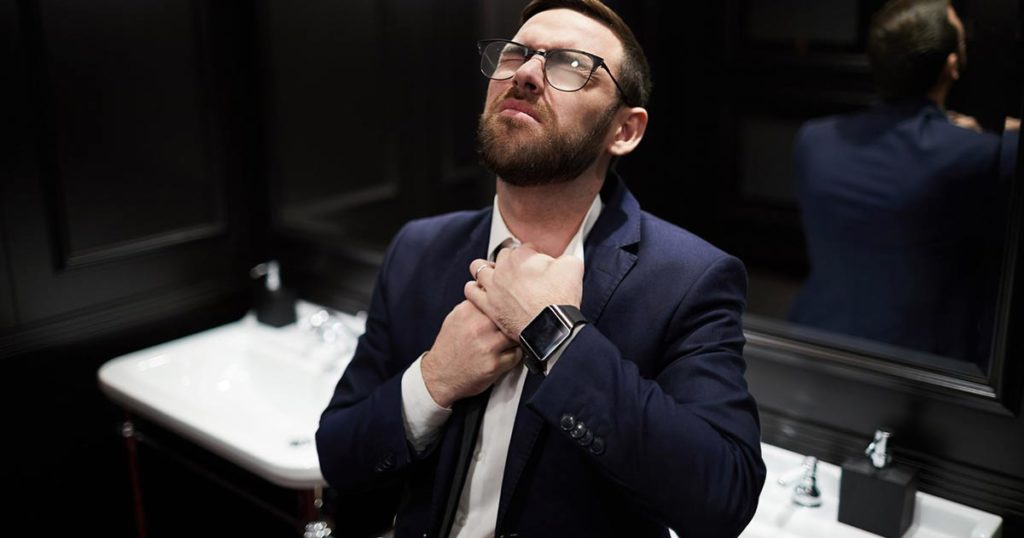 Adult man with glasses in a restroom clutching his chest from an asthma attack. He is wearing a dark suit and has a look of struggle on his face.