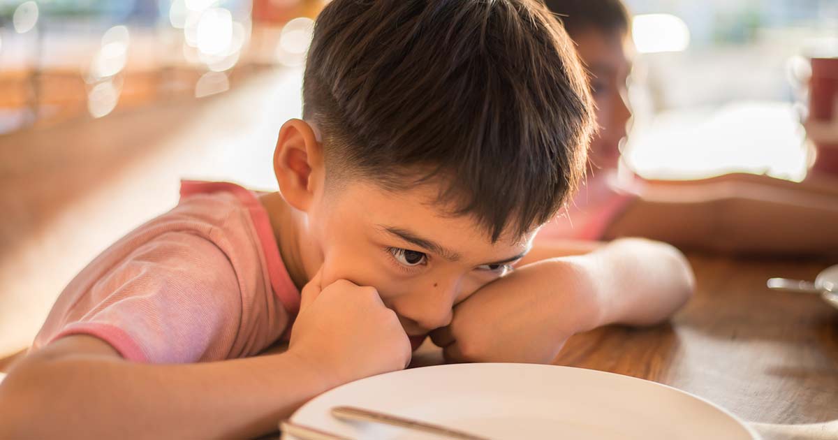 Tween boy sitting at a lunchtable with an empty dish next to him. His hands are on his face in a stressed mood.