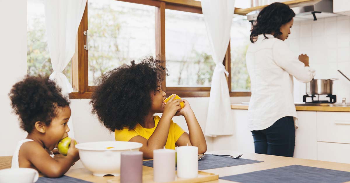 Mother at the kitchen stove cooking, while two tween girls sit at the table looking in her direction.