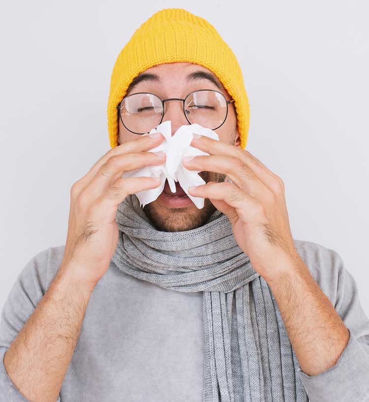 Photo of a young man in a yellow hat and grey shirt. He is holding a tissue to his nose as he