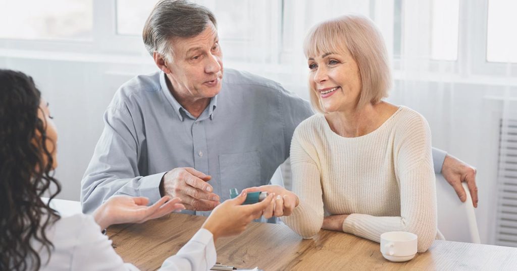 Golden years couple sitting at a desk opposite a doctor. The doctor is handing a woman a fast acting albuterol inhaler as a trial. She is explaining about her new asthma diagnosis and how to manage her symptoms.