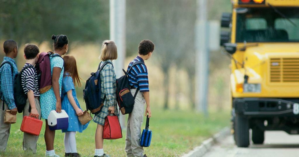 Children in a single file line at the curb as the yellow schoolbus pulls up to pick them up.