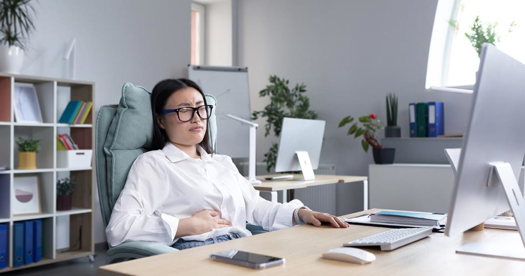 Woman sitting at her desk feeling nauseated and holding her belly.