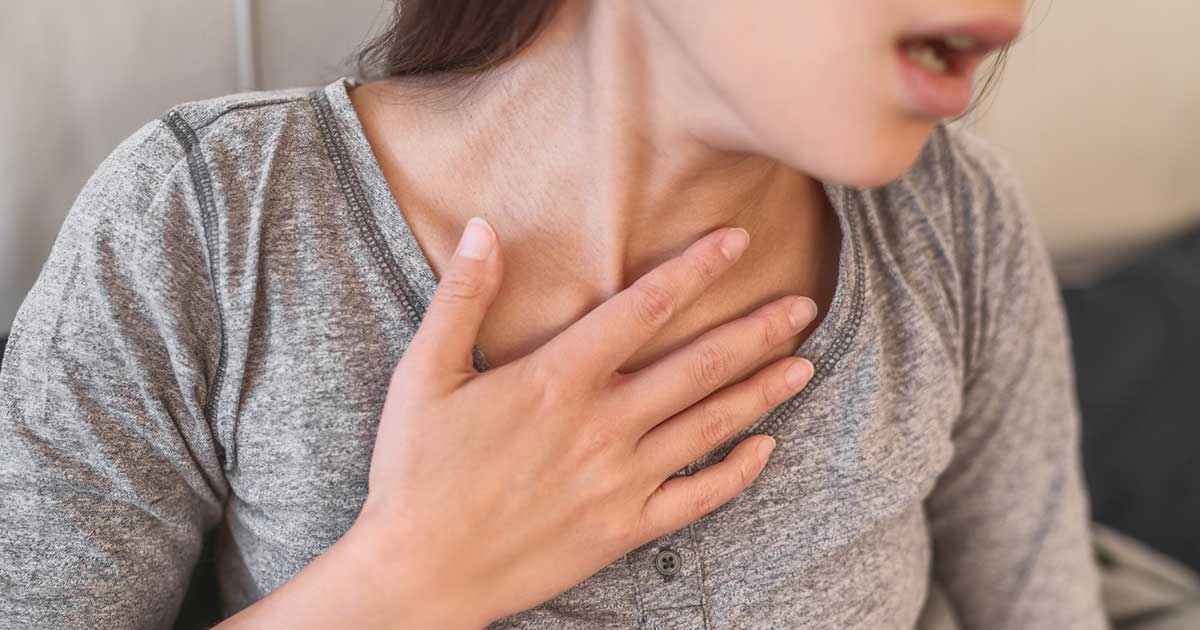 Young adult woman in grey shirt holding her hand to her throat and struggling to breathe because she