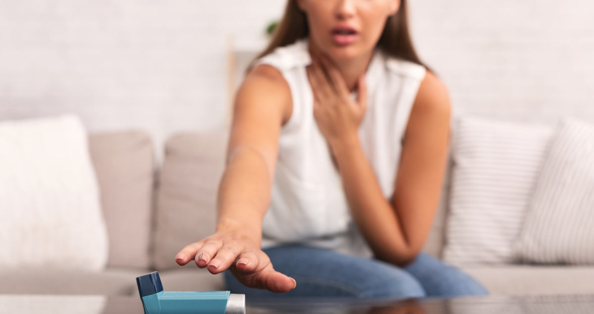 Young adult woman sitting on a couch. She is holding her neck are with one hand and reaching to grab her asthma inhaler with the other hand.