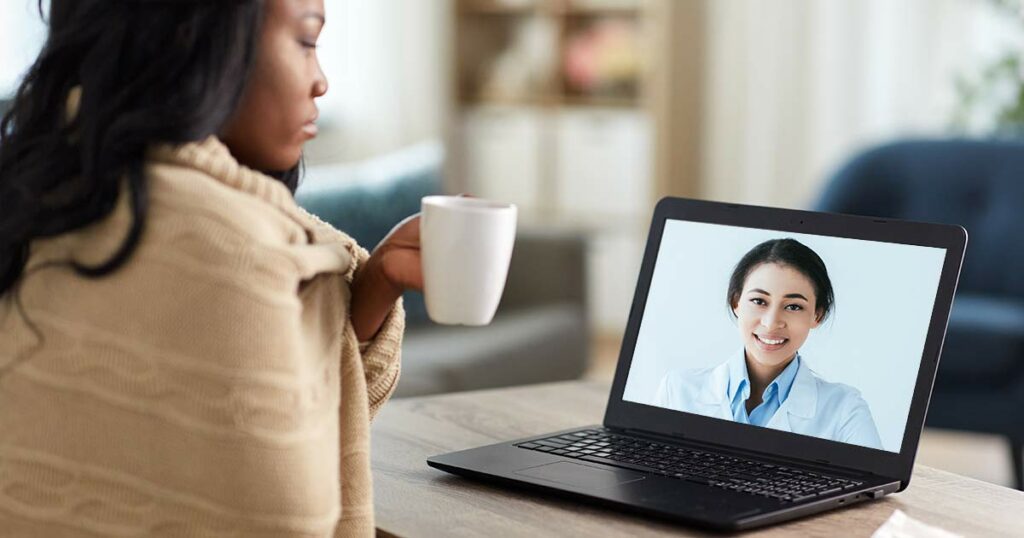 Woman wrapped in blanket, drinking tea, while using a laptop to see an asthma coach online.