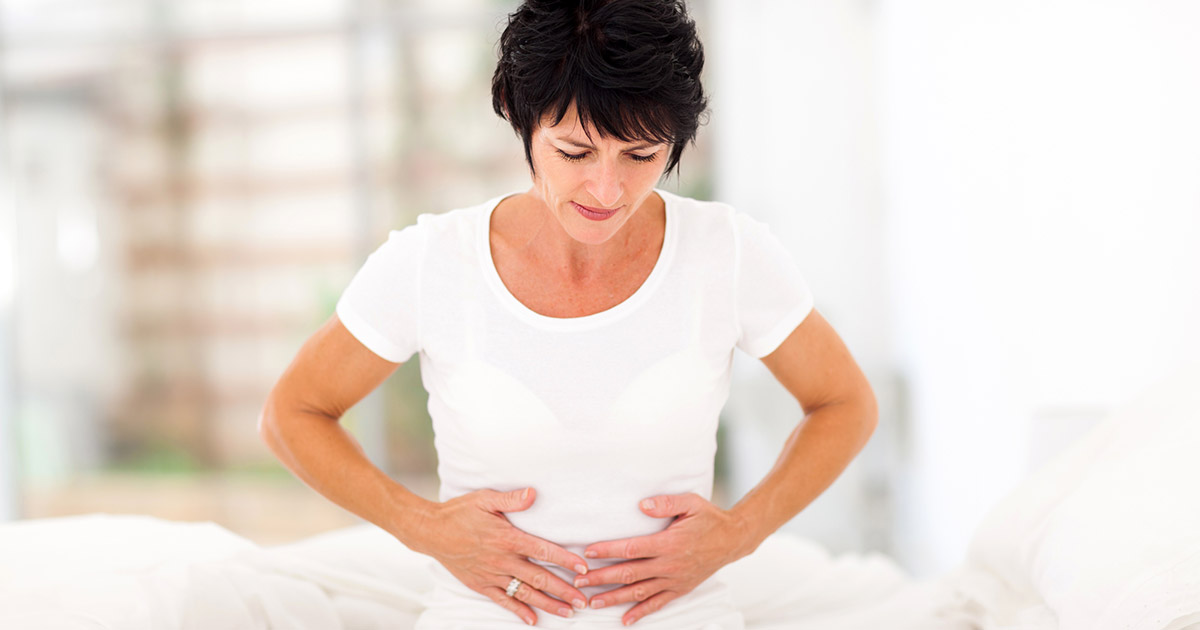Middle aged woman sitting on her bed holding her abdomen in discomfort.