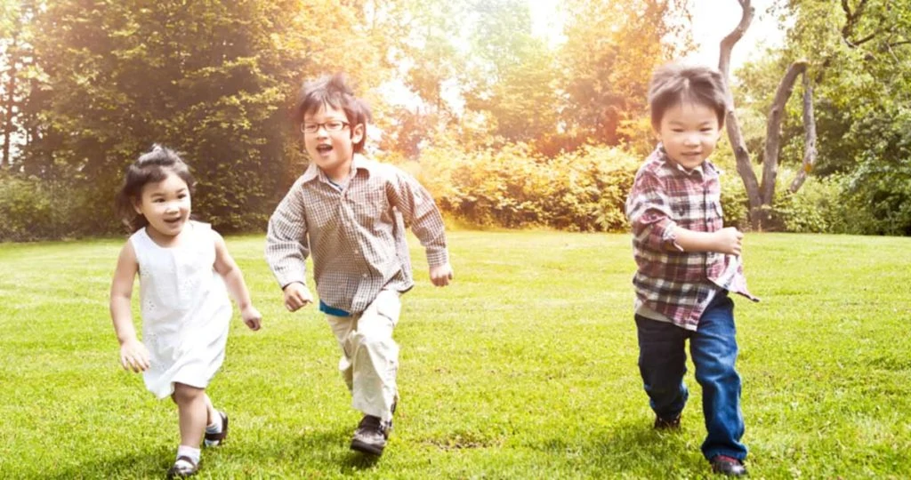 Three small children running in an open field with trees in the background.