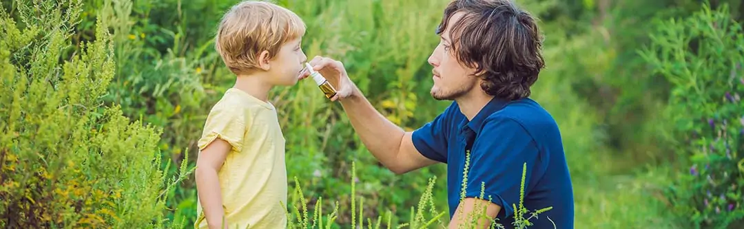 Father giving son antihistamine nasal spray while in a field of grass, bushes, and trees.