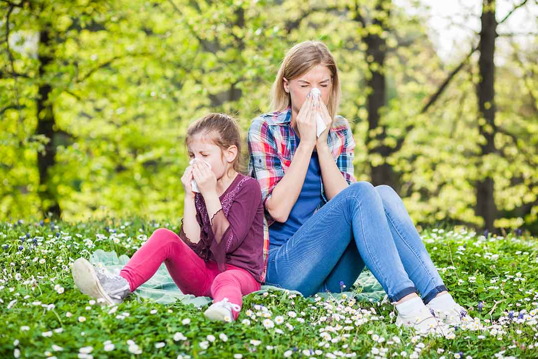 Mother and daughter each blowing their nose with tissues while sitting on grass. There are trees in the background.